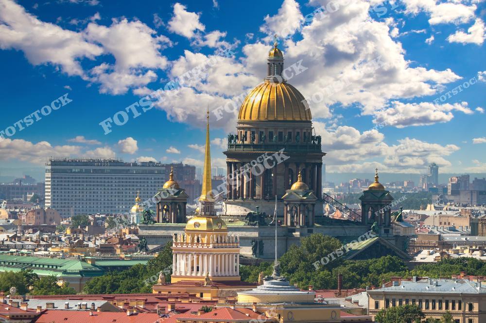 На изображении представлен знаменитый Исаакиевский собор в Санкт-Петербурге. Мощный золотой купол собора ярко выделяется на фоне синего неба и облаков. Вокруг расположены различные здания, зелёные деревья и другие более маленькие купола собора. Вид с высоты птичьего полёта демонстрирует разнообразие и красоту достопримечательностей северной столицы России.